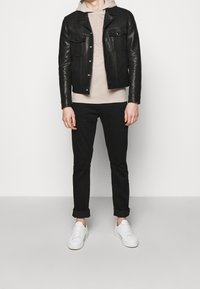 Svart läderjacka med två bröstfickor, beige hoodie under, svarta uppvikta byxor och vita sneakers. Enkelt design med kontrasterande färger.