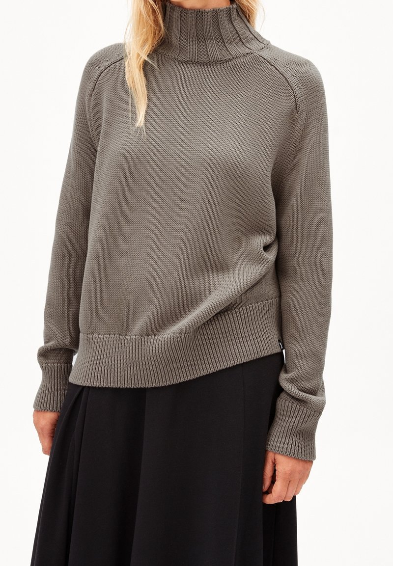 Pull en maille grise avec un col montant côtelé et des poignets côtelés. Présente une coupe décontractée et une surface texturée. Porté avec une jupe noire.