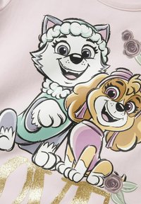 Jasnoróżowy materiał zdobią rysunkowe psy, z czego jeden ma puszystą sierść i niebiesko-zielony strój, a drugi ma złotą sierść z różowymi akcentami; poniżej złoty tekst.