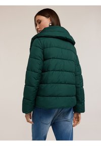 Zelená péřová bunda s kapucí, s horizontálně prošívanými sekcemi, spárovaná s modrými džínami. Hladký povrch, volný střih.