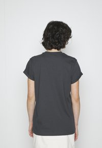 Tamsiai pilkos spalvos medvilninė marškinėliai trumpomis rankovėmis, apvaliu iškirpimu ir suktomis rankovėmis, matomi iš nugaros. Lygus paviršius, minimalus dizainas.