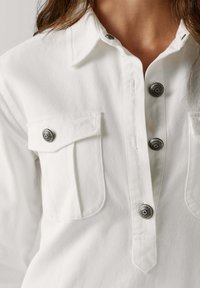 La camicia bianca presenta un collo a camicia, bottoni argentati e due tasche sul petto con tessuto strutturato.