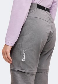 Personne portant un pantalon gris à zip avec le texte "TERREX" sur le côté et une chemise à manches longues violet clair, vue de la taille vers le bas.