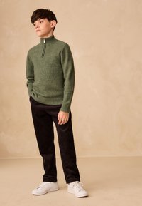 Grøn strikket sweater med høj krave og lynlås, kombineret med sorte fløjlsbukser og hvide sneakers. Simpelt, tekstureret design.