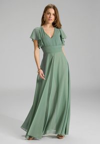 Lang, vloeiende jurk in een zachte groene kleur. Heeft gerimpelde korte mouwen en een aansluitende taille. Gemaakt van lichtgewicht, doorschijnende stof.