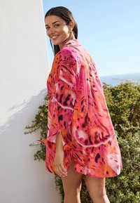 Chemise rose vif et orange à coupe ample, présentant un imprimé animal audacieux et des manches fenduess. La texture semble légère et aérée.