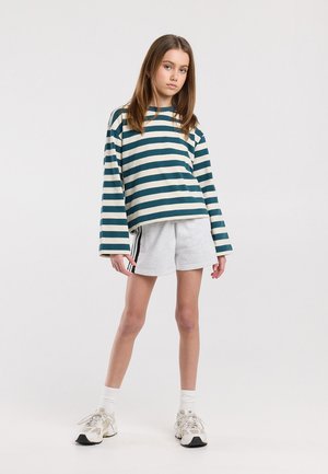 Tienermeisje met een blauwgroen-wit gestreept shirt met lange mouwen, grijze korte broek met zwarte strepen, witte sokken en witte sneakers, staand tegen een witte achtergrond.