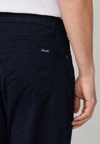 Pantalones azul marino con detalle en el bolsillo trasero, que presenta una pequeña etiqueta de marca blanca y azul. La tela es suave con una textura fina.