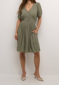 Groene, kortarmige jurk met een V-hals, knoopsluiting aan de voorkant, textuurstof en een geplooide rok. Bevat zijzakken. Gekocht met beige schoenen.