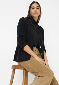 Cardigan nero con una vestibilità rilassata, texture a maglia e grandi bottoni; abbinato a pantaloni di velluto a coste beige, seduto su uno sgabello di legno.