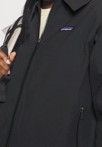 Černá bunda s vysokým límcem, zipovým zapínáním a bočními kapsami. Logo Patagonia v purpurové a bílé barvě na levé části hrudi.