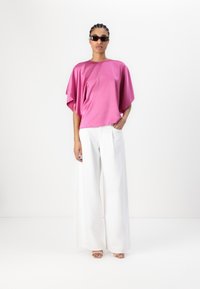 Rosa oversized topp laget av glatt stoff, kombinert med vide hvite bukser. Modellen har på seg mørke solbriller og rosa høye sandaler.