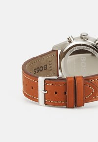 Orologio con cinturino in pelle marrone, dotato di cassa in acciaio inossidabile argentato, cuciture a contrasto e fondo resistente all'acqua con marchio impresso.