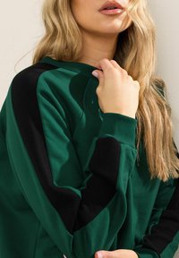 Felpa verde con dettagli neri, caratterizzata da un collo a giro e polsini a coste. Tessuto morbido, vestibilità rilassata e dettagli nella cucitura delle spalle.