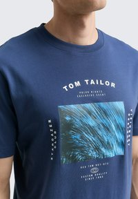 Granatowa bawełniana koszulka z graficznym nadrukiem wirujących niebieskich linii, z białymi elementami tekstowymi i okrągłym dekoltem.