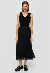 Zwarte mouwloze top met een V-hals, gecombineerd met een geplooide zwarte maxi rok. De rok heeft een vloeiende textuur. Draagt zwarte instappers.