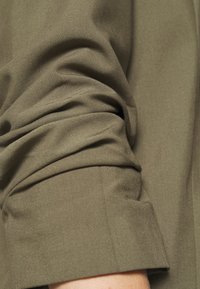 Chemise à manches longues vert olive avec un revers de manche retroussé, présentant un tissu lisse et texturé ainsi qu'un détail de couture subtil sur la manche.