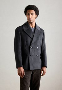 Donkergrijze dubbele-breasted wollen peacoat met een brede kraag, voorzien van twee zijzakken en zwarte knopen, gedragen over een grijs shirt.