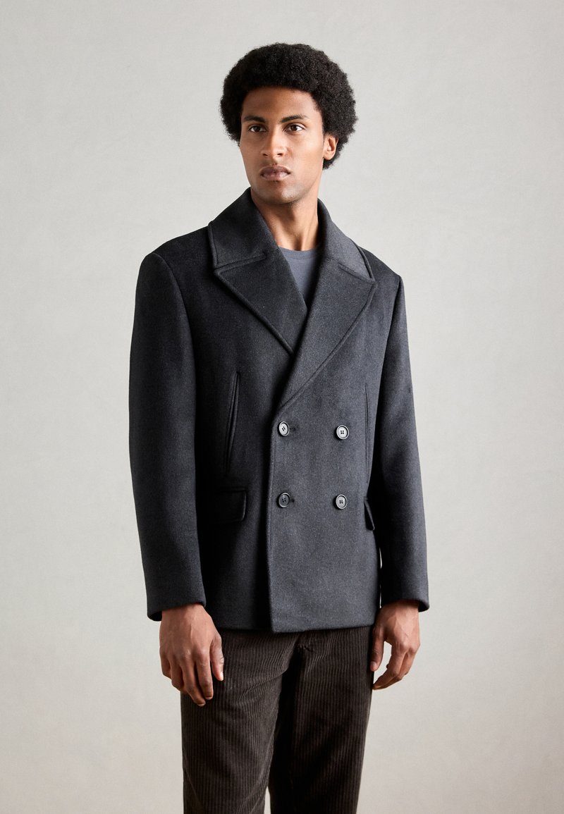 Donkergrijze dubbele-breasted wollen peacoat met een brede kraag, voorzien van twee zijzakken en zwarte knopen, gedragen over een grijs shirt.