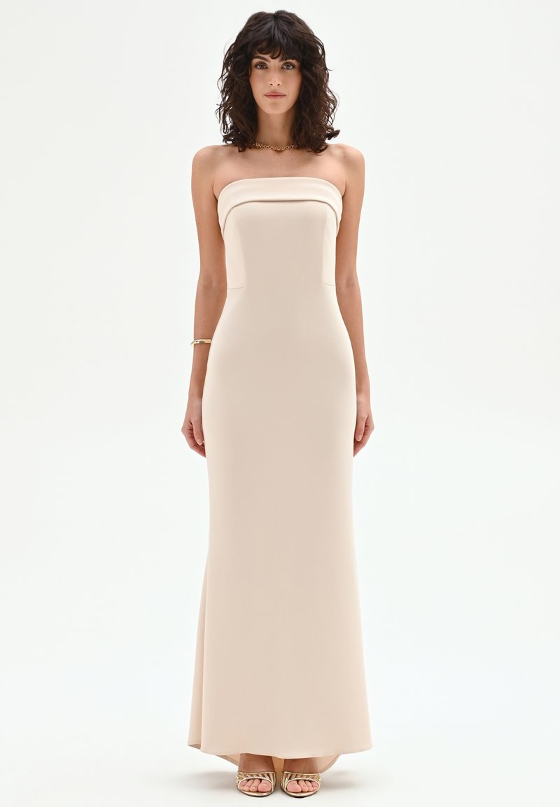 Vrouw met krullend donker haar, gekleed in een strapless, vloerlang beige galajurk en gouden sandalen, staand tegen een effen witte achtergrond.