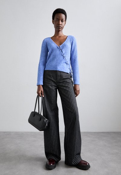Pulover ribbed de culoare albastru deschis, cu decolteu în V și detaliu cu șireturi, asortat cu pantaloni largi cu dungi negre și albe și o geantă neagră.