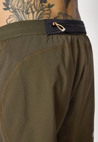 Olive green athletic shorts with an elastic waistband, featuring a black zippered pocket and orange drawstring detail. Smooth texture.