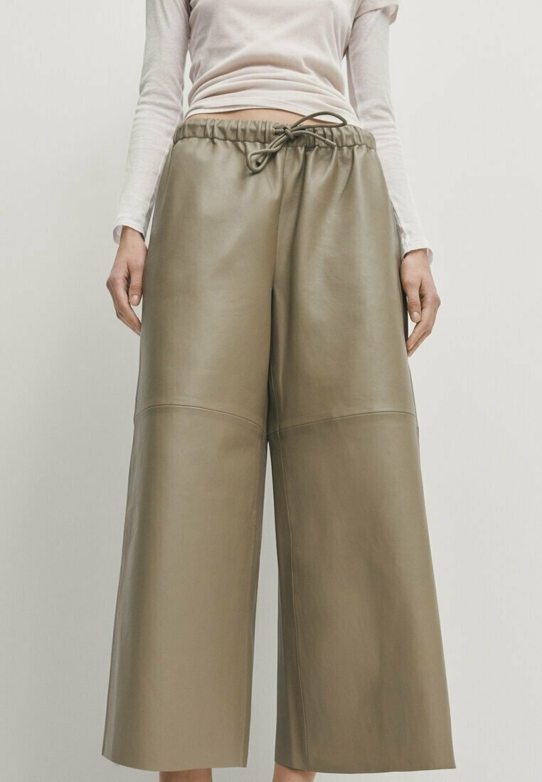 Femme portant un pantalon en cuir vert olive taille haute à jambes larges, avec une taille élastique et un cordon, associé à un haut blanc à manches longues.