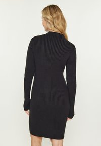 Rochie din tricot neagră, cu guler înalt și mâneci lungi, siluetă mulată și lungime până la mijlocul coapsei. Detalii din material texturat vizibile.