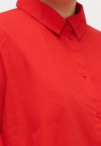 Primo piano di un colletto di camicia rossa e della parte superiore del petto di una persona su uno sfondo neutro.