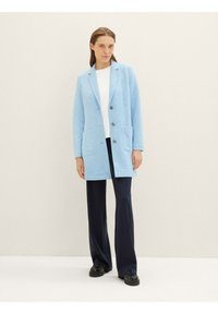 Light blue long blazer in textured fabric, featuring a notched collar, two front pockets, three buttons, and paired with dark trousers.