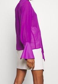 Lila transparente Bluse mit langen, glockenförmigen Ärmeln und Knopfdetails an den Bündchen, kombiniert mit einem hellen Rock. Einfaches, elegantes Design.