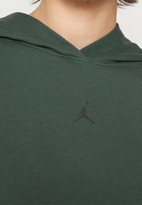 Gros plan d'une personne portant un sweat à capuche vert avec un petit logo Jordan noir centré sur la poitrine.