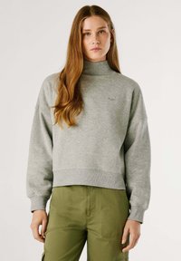 Sudadera gris, recortada, con cuello alto y puños acanalados. Presenta un pequeño logo en el pecho. Lleva puesta con pantalones verde oliva.