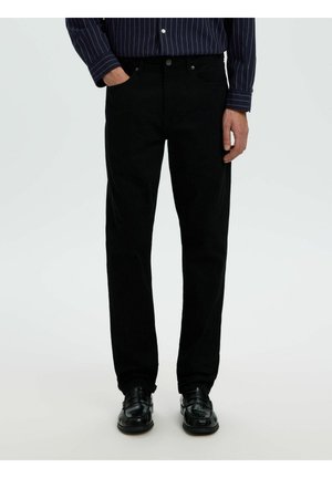 Homme portant un pantalon noir, des chaussures de ville noires brillantes et une chemise bleu foncé à manches longues avec des rayures verticales blanches, debout avec la main dans la poche.