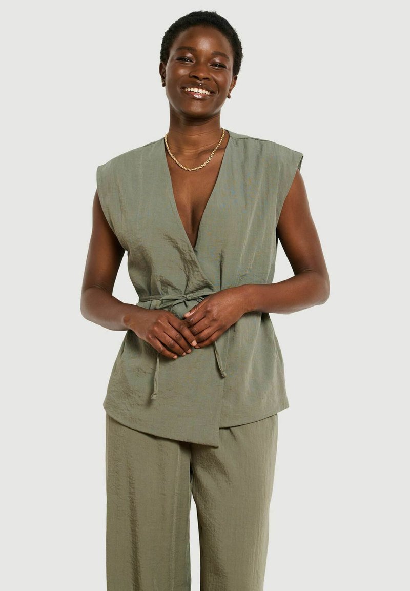 Donna sorridente che indossa un top incrociato senza maniche verde oliva chiaro con cintura e pantaloni ampi abbinati, in piedi con le mani incrociate.