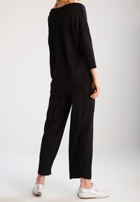 Svart, lös passande topp med trekvartslånga ärmar; kombinerad med svarta, vida byxor och silverfärgade sneakers. Slät textur genom hela plagget.