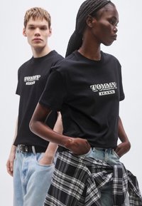 Fekete pamut pólók a "TOMMY JEANS" logóval, amelyeket modellek viselnek különféle farmernadrágokkal és kockás inggel, amelyet a derék köré kötöttek.