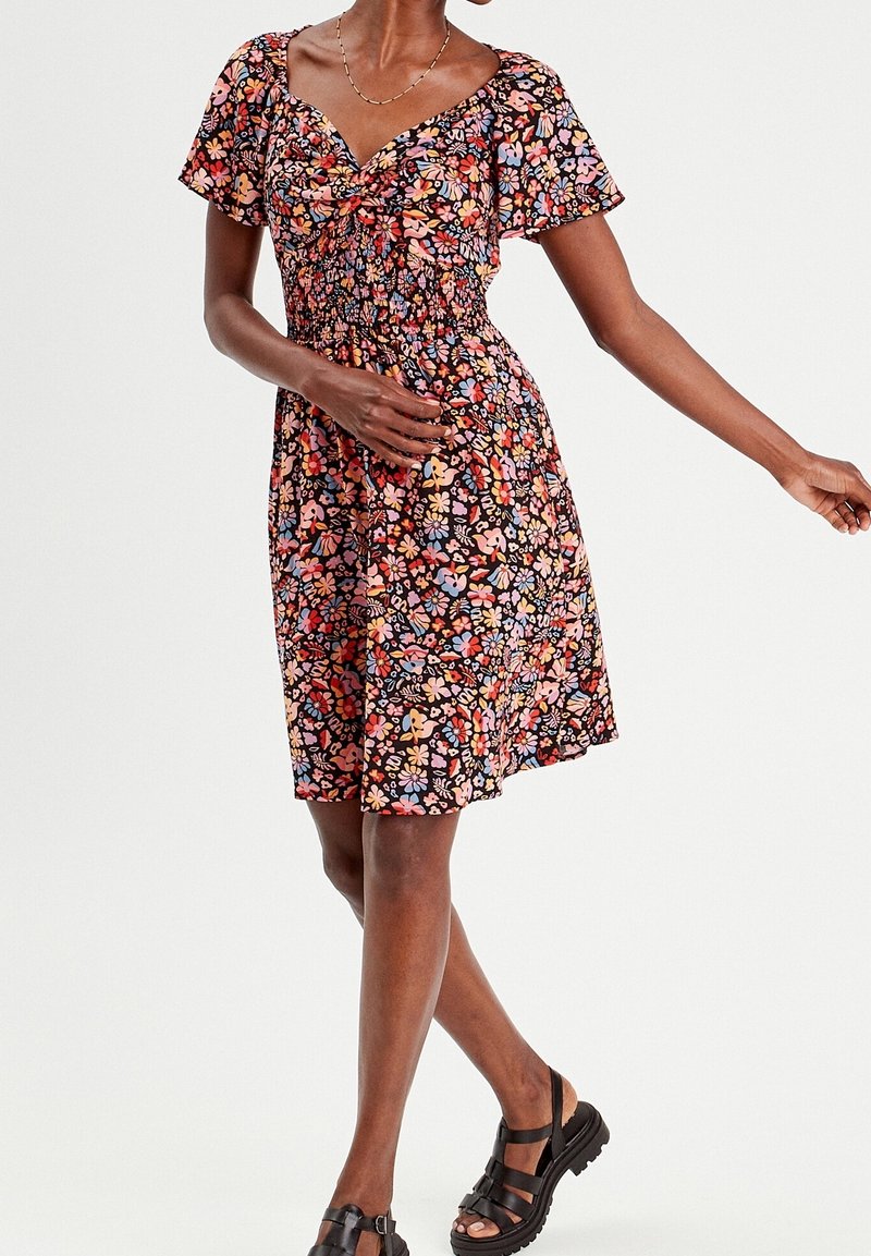 Femme portant une robe mi-longue à imprimé floral multicolore, manches courtes et sandales noires épaisses sur fond blanc.