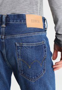 Blå denimjeans med en läder EDWIN-logotyp, med gul sömnad och en rundad fickdesign på baksidan.
