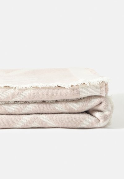 Couverture beige douce pliée avec un motif zigzag subtil et des bords à franges sur un fond blanc uni.