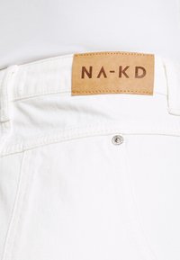 Bílé džíny s hnědou koženou cedulkou s nápisem "NA-KD". Obsahují kovové zapínání na knoflík a viditelné švy podél pasu.