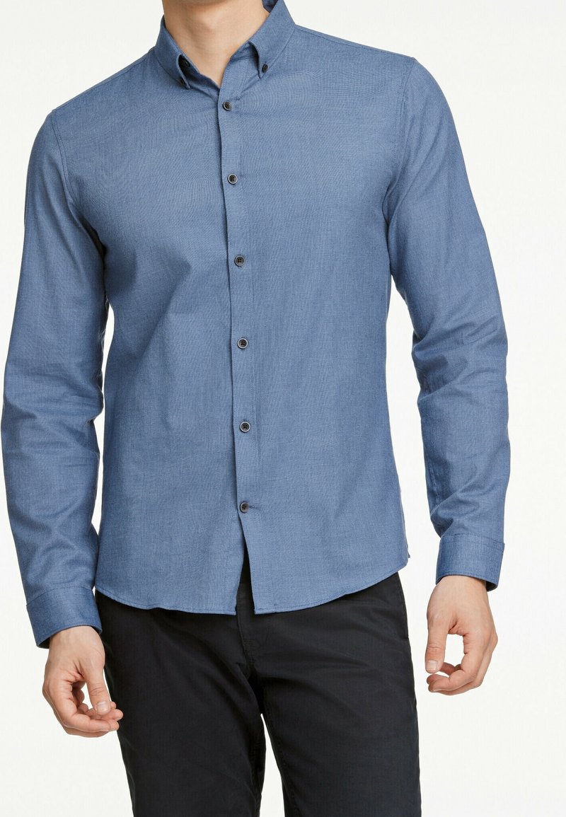Camicia button-up azzurra chiaro con maniche lunghe, colletto, bottoni neri e una texture sottile. Indossata con pantaloni scuri.
