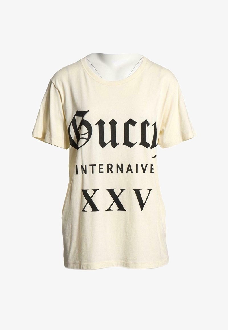T-shirt crème à manches courtes avec un texte noir ""Gucci Internaive XXV"" en grandes lettres stylisées sur le devant.