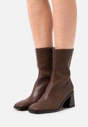 Botas de cuero marrón hasta la mitad de la pantorrilla con tacones de bloque, usadas sobre piernas desnudas frente a un fondo blanco liso.