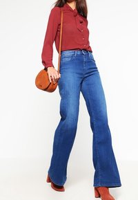 Blouse rouge côtelée avec détail à nouer, assortie à un jean évasé bleu et un sac bandoulière marron. Le mannequin porte des bottines rouges à talons.