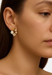 Ohrringe in Goldfarbe mit einem Schmetterlingsdesign, einer weißen Perlenperle und Blütenakzenten mit klaren Steinen, vor einem neutralen Hautton.