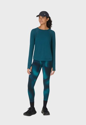 Vrouw die een donker teal gekleurde top met lange mouwen draagt, teal en zwart patroonleggings, zwarte sneakers en een zwarte pet, staande op een witte achtergrond.