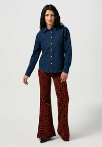 Jeansskjorta med spetsig yoke-design och vita knappar, i kombination med utsvängda röda byxor med ett geometriskt mönster i mörkare nyanser.