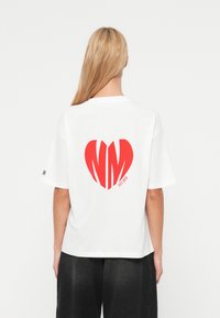 Λευκό βαμβακερό μπλουζάκι με μεγάλο κόκκινο καρδιά αποτύπωμα που περιλαμβάνει τα γράμματα "NM" στο εσωτερικό και "EST. 2023" από κάτω. Άνετη εφαρμογή με κοντά μανίκια.