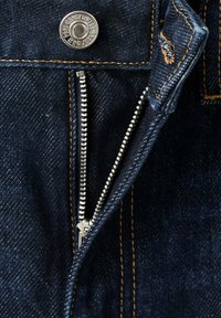 Los pantalones de mezclilla oscura presentan un acento de costura dorada, un botón de metal en la cintura y una cremallera plateada parcialmente abierta en la parte delantera.